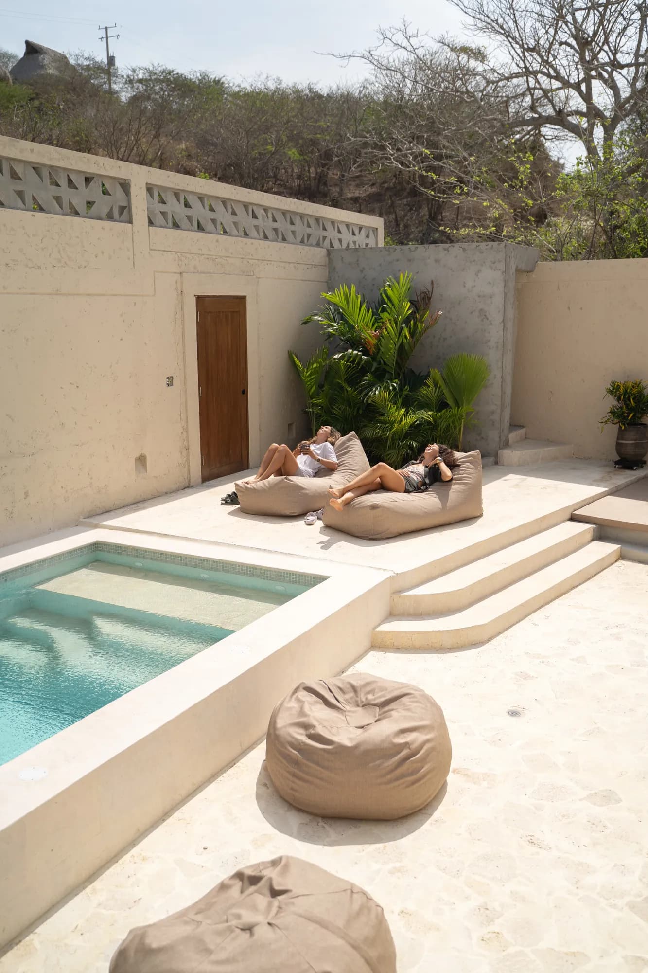 Huespedes descansando en puffs junto a la piscina rectangular con escalones de piedra y colina de fondo