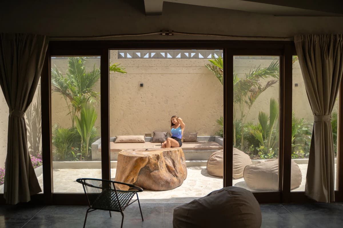 Area de piscina en patio con puffs, plantas tropicales, piso de piedra y persona caminando