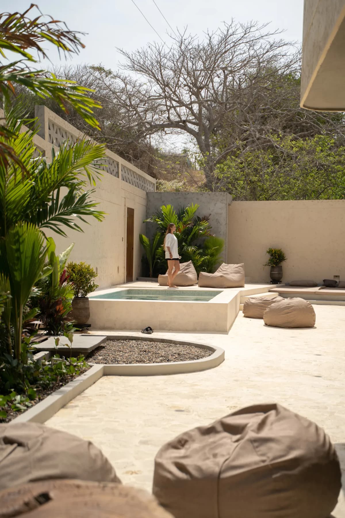 Vista desde la cocina del patio con piscina, plantas tropicales, puffs y maceta decorativa de piedra