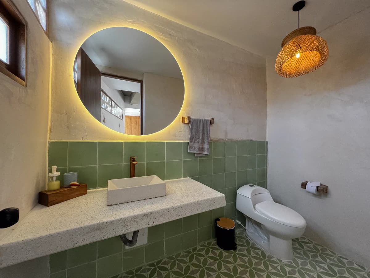 Bano con salpicadero de azulejo verde, espejo redondo retroiluminado, lampara tejida y piso con patron