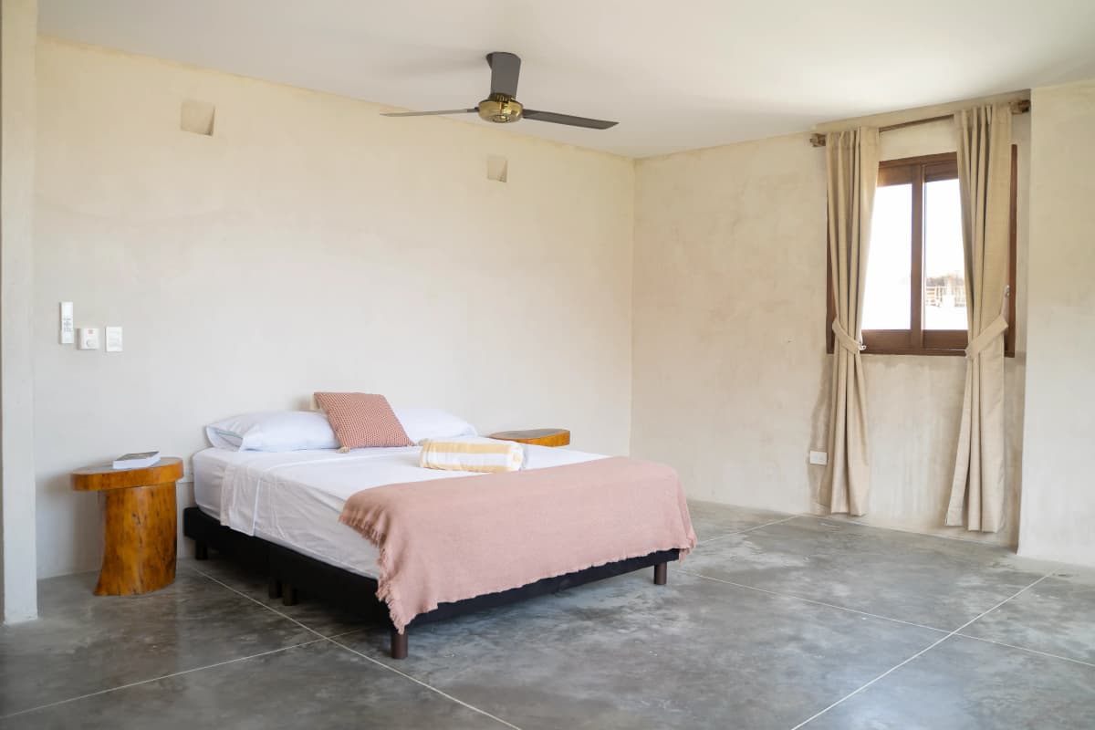 Habitacion con cama king, manta terracota, mesa de noche de madera, ventilador de techo y ventana con cortina