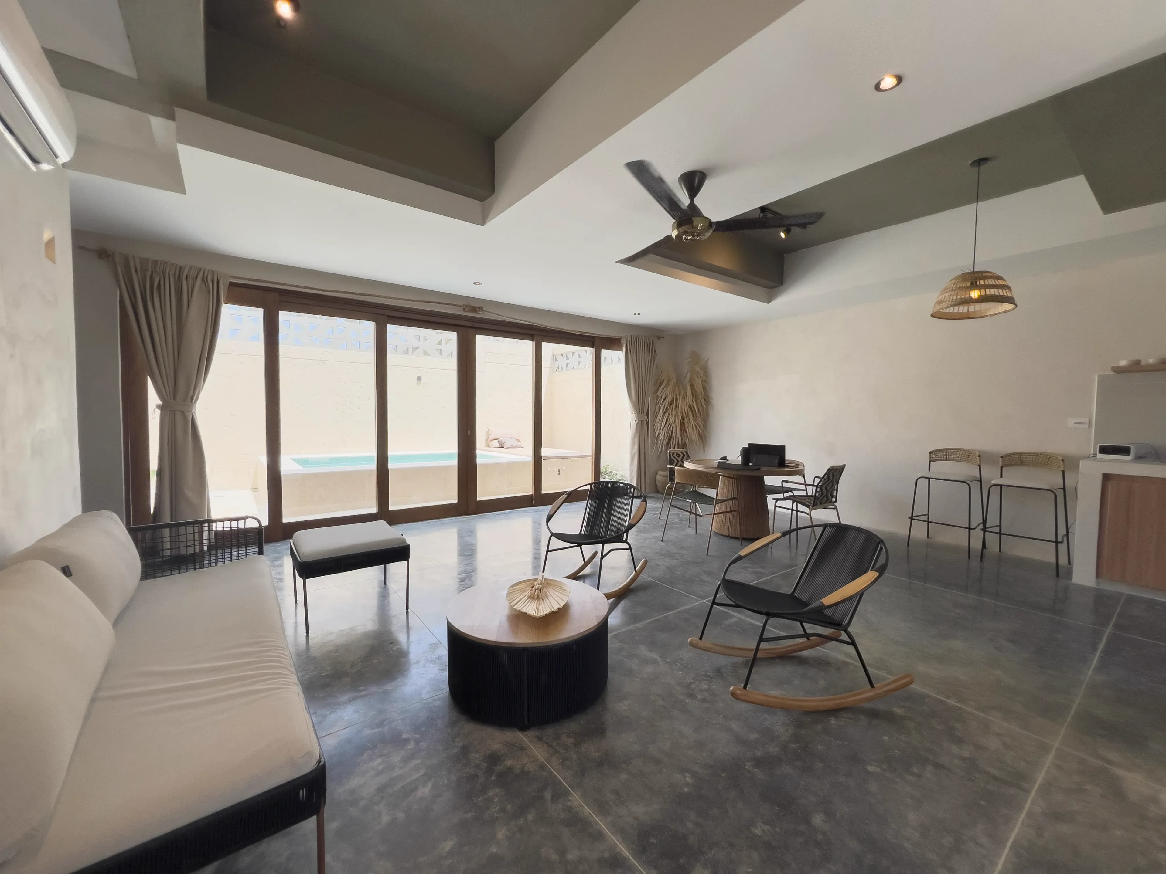 Sala abierta con sofa, mecedoras, taburetes de barra y puertas de vidrio hacia el area de piscina