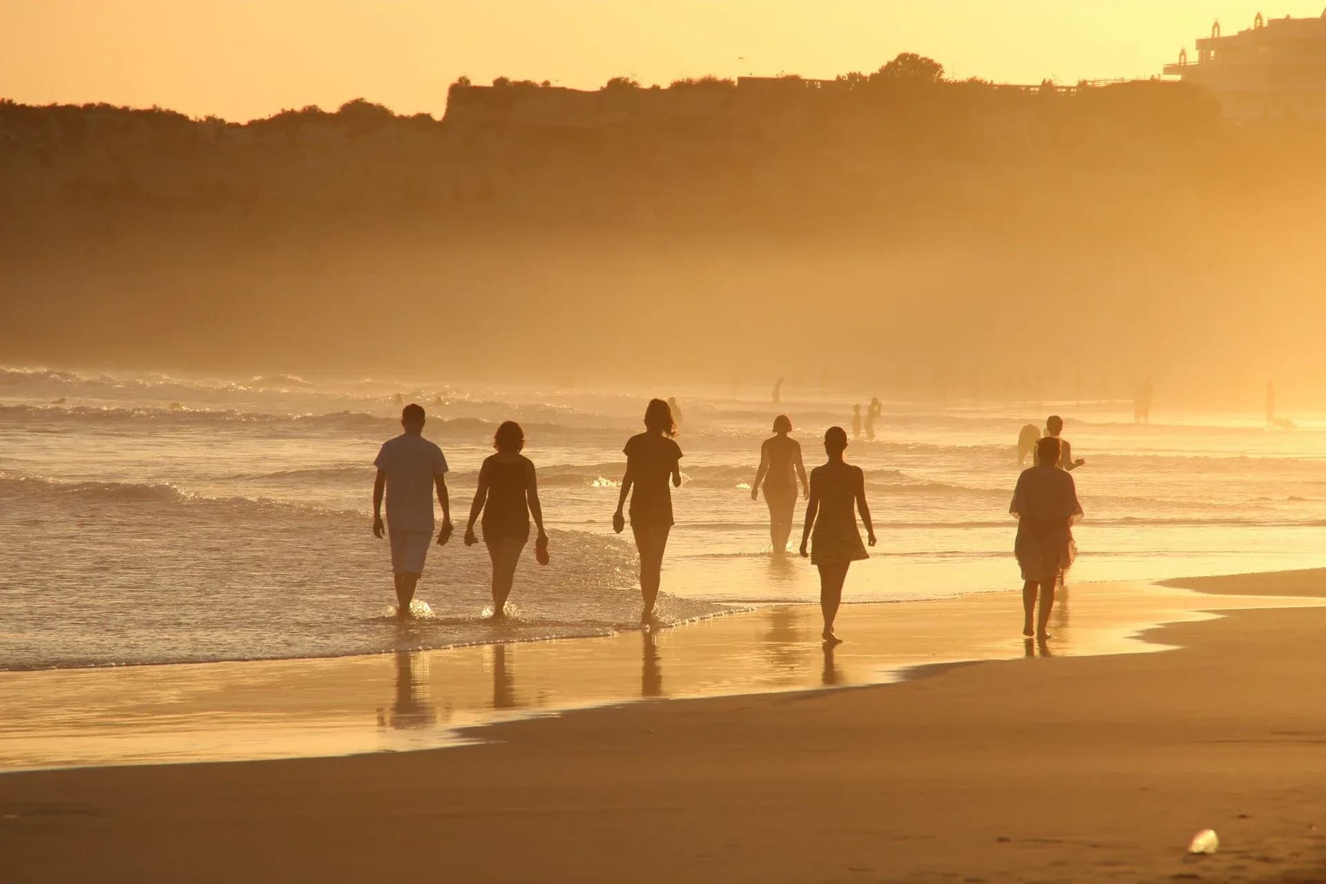 Siluetas de personas caminando por la playa al atardecer dorado con olas y colinas brumosas
