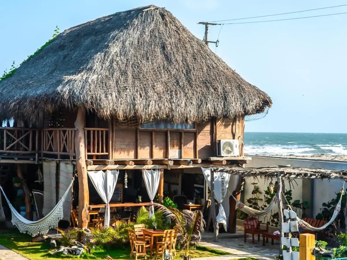 Hostal frente al mar con techo de paja, hamacas, muebles de madera y olas del mar al fondo