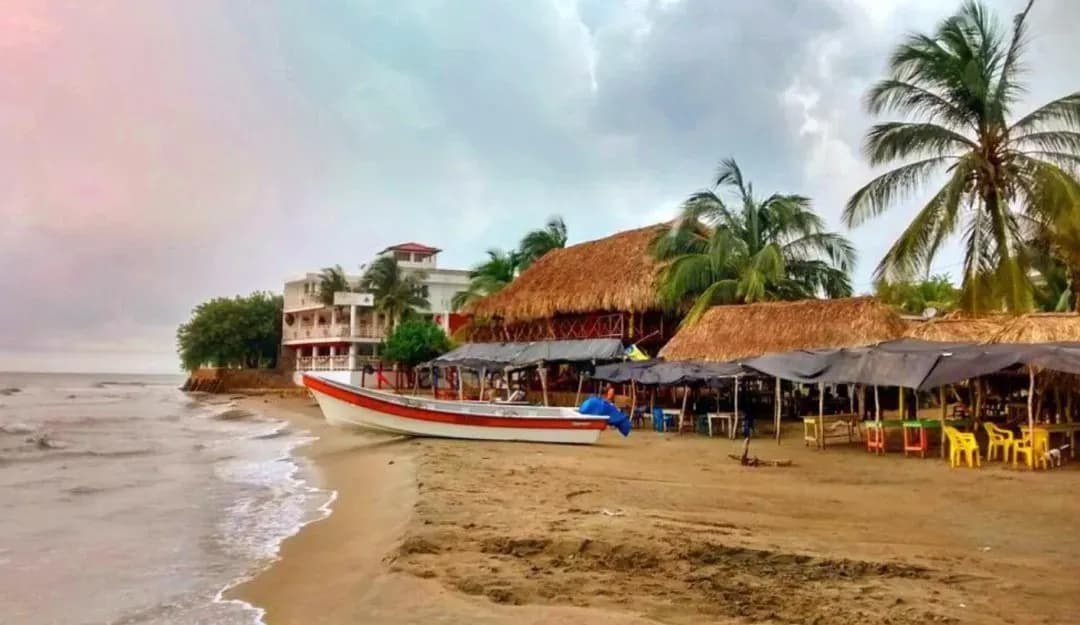 Bote de pesca colorido en la playa con restaurantes de techo de paja y palmeras bajo cielo nublado