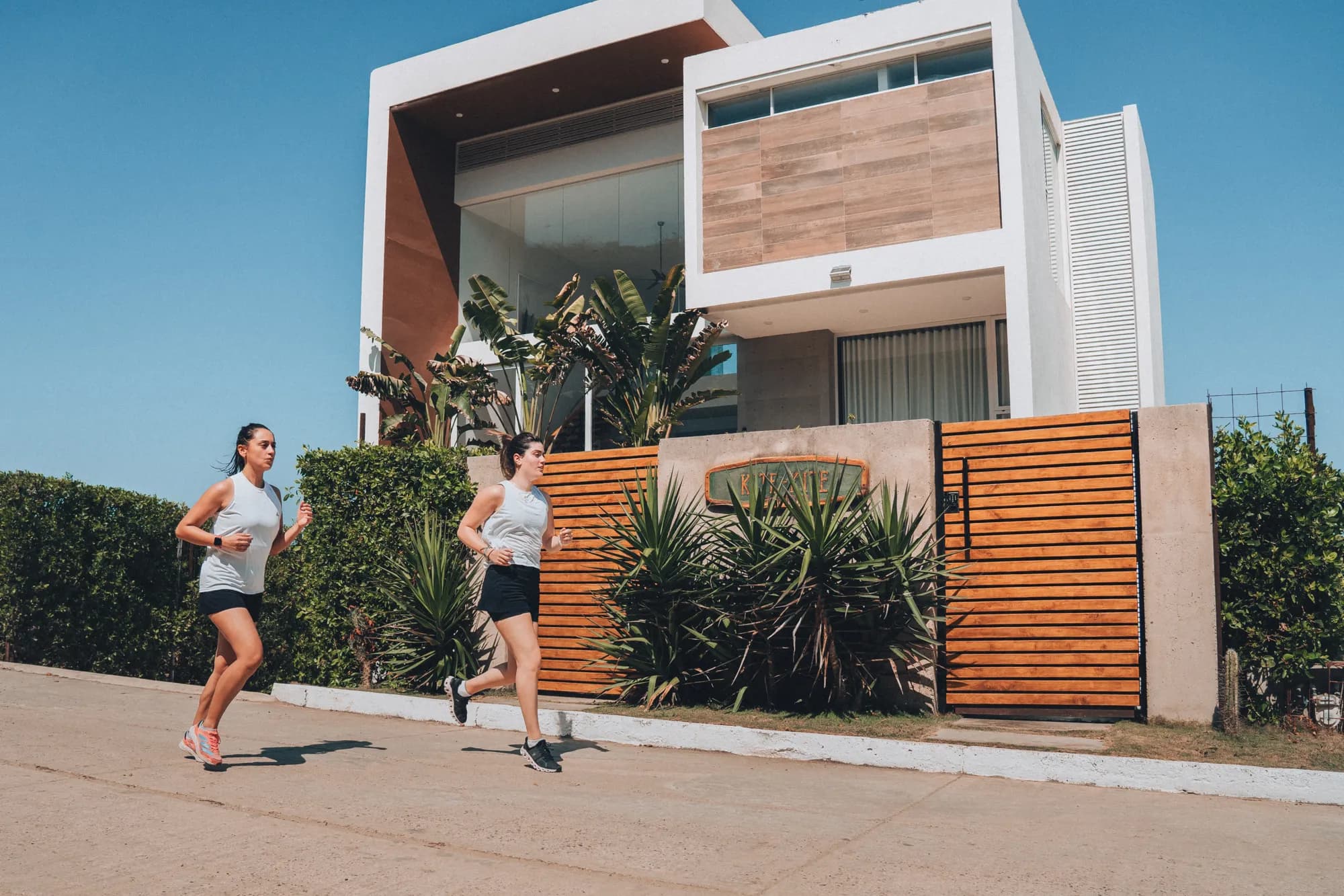 Dos mujeres corriendo frente a una casa de playa moderna con plantas tropicales en una manana soleada