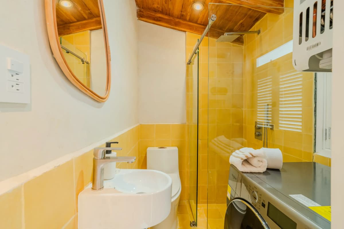 Bano compacto con azulejos amarillos brillantes, espejo redondo, ducha de vidrio y lavadora