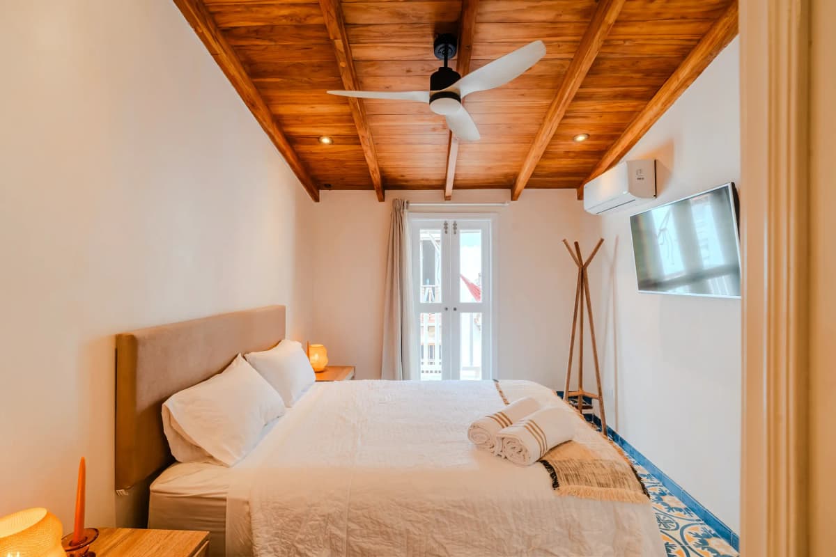 Habitacion con cama queen, techo de vigas de madera, ventilador de techo, puerta al balcon y TV en la pared